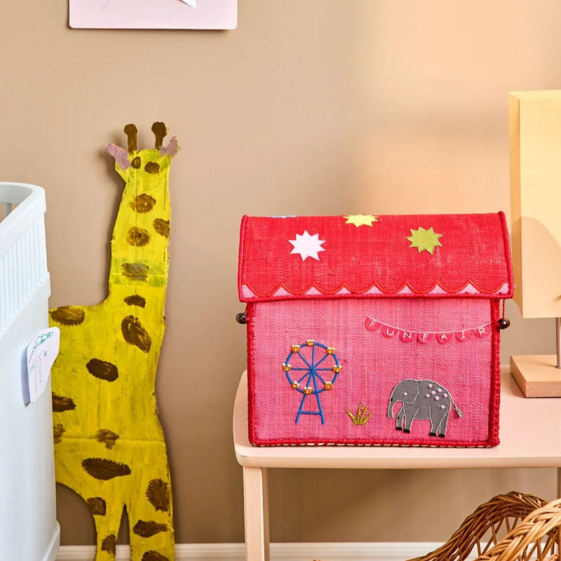 En opbevaringskurv formet som et hus i raffia materiale. Huset er pink med rødt tag og er udsmykket med en elefant, pariserhjul og andre sjove Fun Fair motiver.
