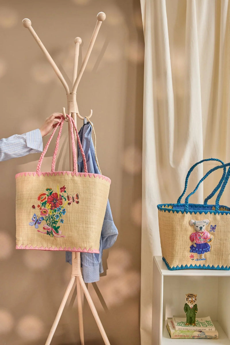 Taske i naturmaterialet raffia. Tasken har en lyserød kant og hank. På midten er der smukke blomster af lyserød, blå, rød og gul med smukke sommerfugle omkring.