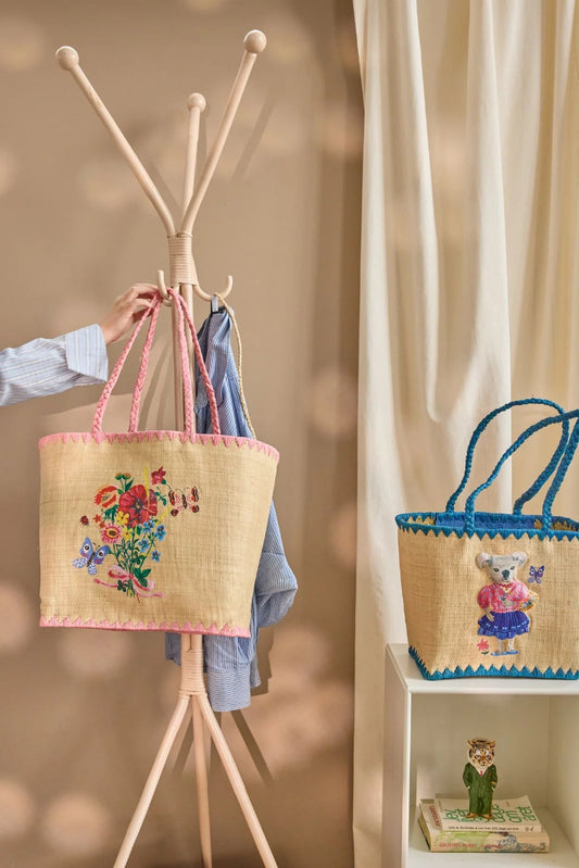 Taske i naturmaterialet raffia. Tasken har en lyserød kant og hank. På midten er der smukke blomster af lyserød, blå, rød og gul med smukke sommerfugle omkring.