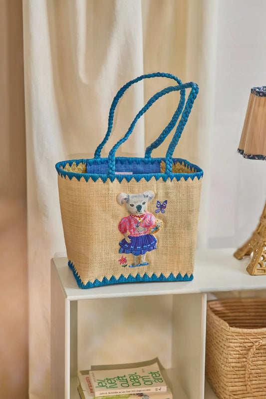 Taske i raffia materiale med blå kant og hank. På midten er motivet af en koala bjørn med tøj på omkringet af sommerfugle og blomster.